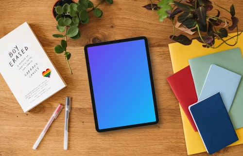 Tablet mockup on a wooden desk with pride enamel pin and colorful notebooks