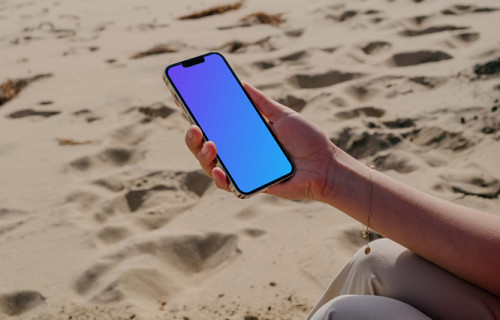 iPhone 13 Pro mockup held by a user seated on sand at the beach