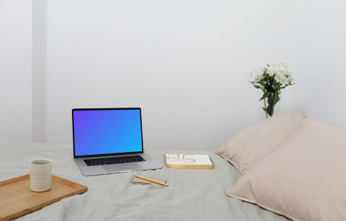 MacBook mockup en una cama con un libro y una bandeja de madera al lado.