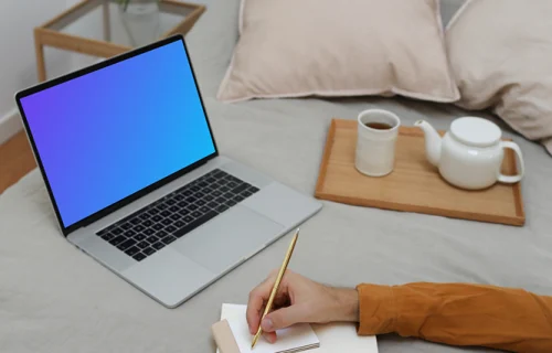MacBook mockup sobre una cama con un usuario escribiendo en un cuaderno
