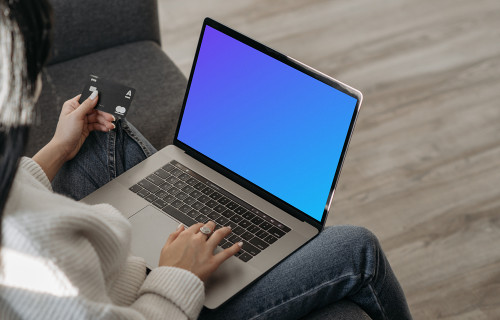 Mockup de una usuaria en un sofá comprando en línea con su MacBook gris y una tarjeta MasterCard en la mano